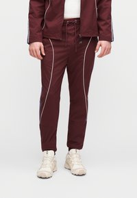 Bordó sportnadrág fehér csíkozással, húzózsinóros derékrésszel és kék oldalsó díszítéssel, krém színű tornacipővel párosítva. Finom anyagú és szűkített szabású.