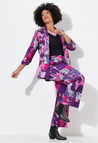 Živý dvoudílný outfit s abstraktními vzory obličejů v růžové a fialové barvě, se fitted černým tričkem a hrotovanými černými kotníkovými botami.