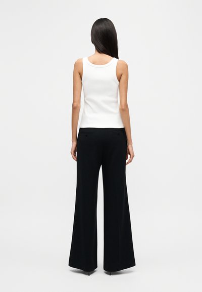 Λευκή ριμπ μπλούζα χωρίς μανίκια συνδυασμένη με μαύρο, φαρδύ παντελόνι ψηλόμεσο με πίσω τσέπες και λεία υφή, σε θέα από πίσω.