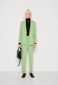 Mintgrüner Blazer und passende taillierte Hose, kombiniert mit einem schwarzen Rollkragenpullover. Schwarze Slipper und eine strukturierte schwarze Handtasche sind ebenfalls enthalten.