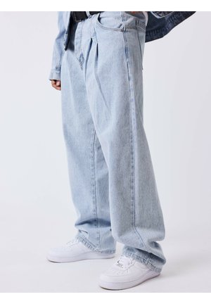 Personne portant un jean bleu clair ample et des baskets blanches, debout devant un fond clair uni.