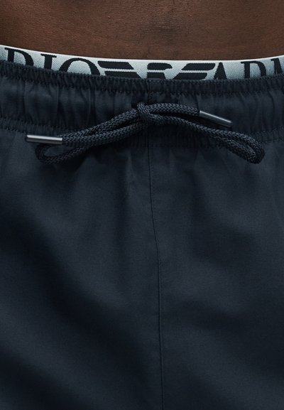 Μαύροι ελαστικοί σορτς με κορδόνι δεμένο σε φιόγκο, που αποκαλύπτει μια λευκή ζώνη μέσης με μερικώς ορατό φαρδύ μαύρο κείμενο.