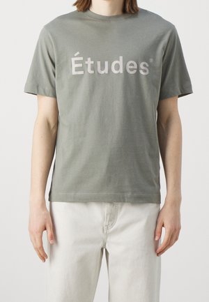 Osoba ubrana w przygaszony zielony t-shirt z krótkim rękawem z napisem "Études" oraz jasne spodnie, stojąca na jednolitym białym tle.