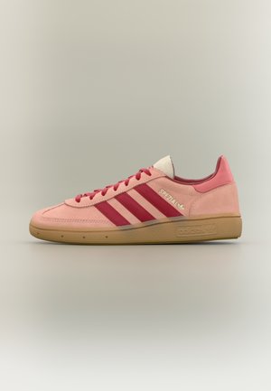 Różowy zamszowy sneaker Adidas z czerwonymi paskami, gumową podeszwą, napisem "Spezial" po boku i czerwonymi sznurowadłami na neutralnym tle.