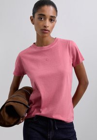 T-shirt à manches courtes, coupe classique, en tissu doux de couleur rose clair avec un détail logo subtil. Le modèle tient un sac en cuir marron.