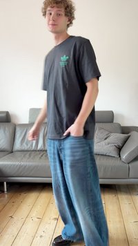 Μαύρο μπλουζάκι με πράσινο λογότυπο, φαρδιά ανοιχτό μπλε τζιν, στέκεται σε ξύλινο πάτωμα δίπλα σε γκρι καναπέ με διακοσμητικά μαξιλάρια.