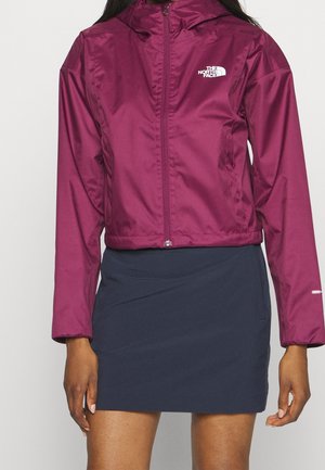 Veste coupe-vent bordeaux avec fermeture éclair et capuche, arborant un logo sur la poitrine et un design court. Associée à une jupe marine.