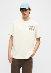 Ung mann iført en beige T-skjorte med «Nature in Motion» og jordklode-design på brystet, blå Nike-caps og brune bukser, stående med en hånd i lommen.