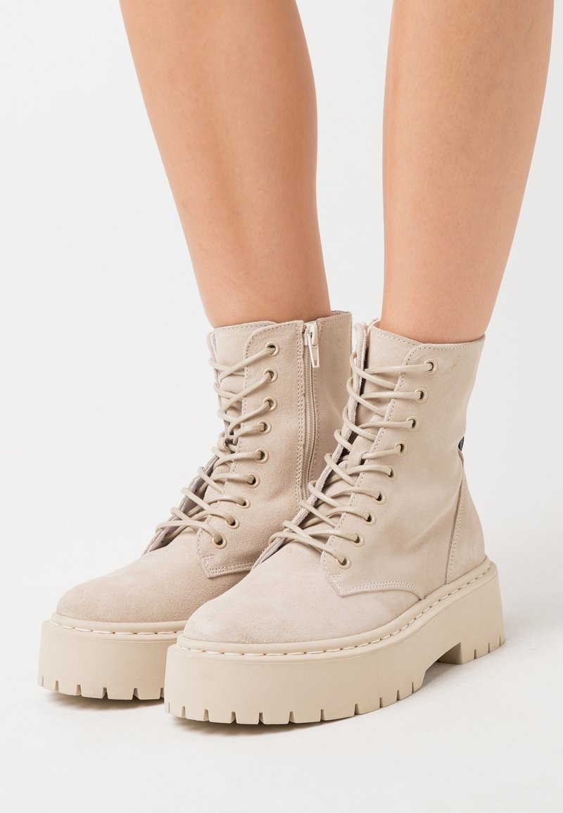 Bottines en daim beige avec une semelle plateforme épaisse. Dotées de lacets et d'une fermeture éclair latérale, elles présentent un matériau texturé et un design robuste.