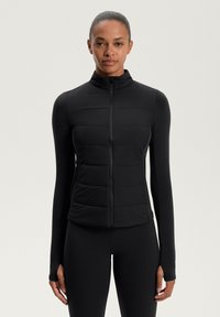 Una donna che indossa un gilet imbottito nero con zip sopra un top a maniche lunghe aderente e leggings neri abbinati, in piedi contro uno sfondo chiaro e uniforme.