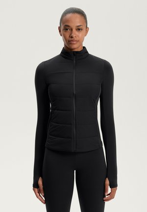Frau in einer schwarzen, gepolsterten, Reißverschlussweste über einem enganliegenden Langarmoberteil und passenden schwarzen Leggings, die vor einem schlichten hellen Hintergrund steht.