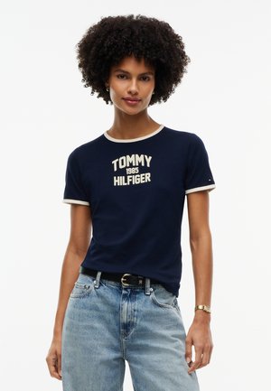 Femme aux cheveux bouclés portant un t-shirt bleu marine Tommy Hilfiger et un jean bleu clair, debout devant un fond blanc.