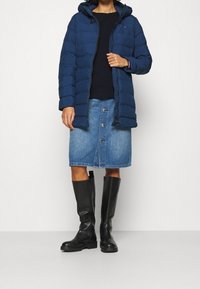 Chaqueta de plumas azul marino con capucha, mangas largas, costuras verticales, cierre de cremallera, llevada sobre un suéter negro y una falda de mezclilla azul, combinada con botas altas negras.