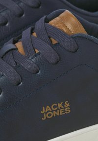 Navy blue sneakers with brown leather accents, textured laces, and gold logo detail. Smooth upper material with contrasting stitching.