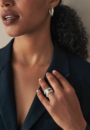 Sølv hoops øreringe og en sølv ring med en struktureret band bæres på en hånd over en mørk blazer, hvilket viser et poleret look.