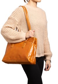 Borsa tote in pelle color cuoio con forma strutturata, doppi manici e cuciture a vista. Portata da una persona con un maglione beige texture.