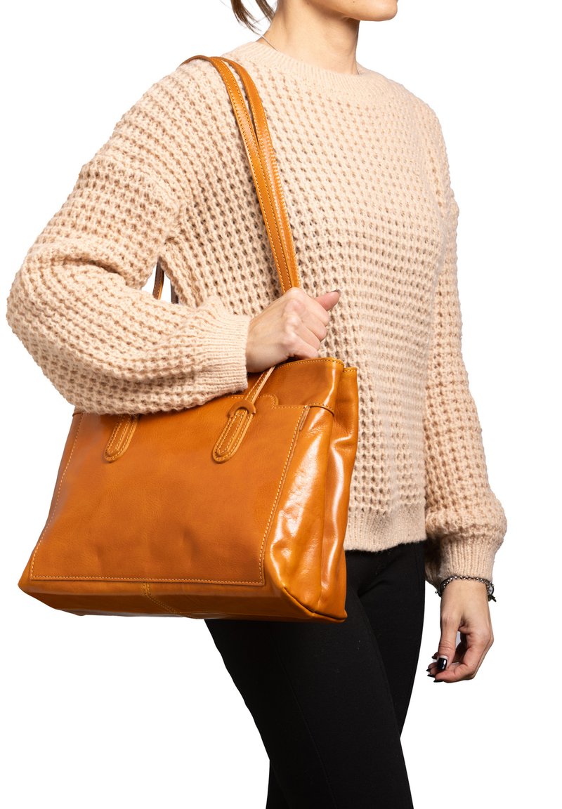 Borsa tote in pelle color cuoio con forma strutturata, doppi manici e cuciture a vista. Portata da una persona con un maglione beige texture.