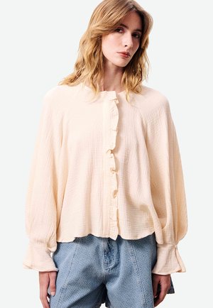 Jonge vrouw die een losse crèmekleurige blouse met ruches en lange mouwen draagt, gecombineerd met lichtblauwe, gestructureerde, high-waisted korte broek, staand tegen een effen achtergrond.