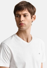 Jonge man met kort bruin haar, die een wit T-shirt met V-hals draagt, kijkt naar links tegen een effen lichte achtergrond.