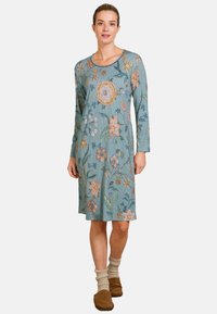 Woman wearing a knee-length, long-sleeve teal dress with multicolored floral patterns, beige socks, and brown slip-on shoes standing.