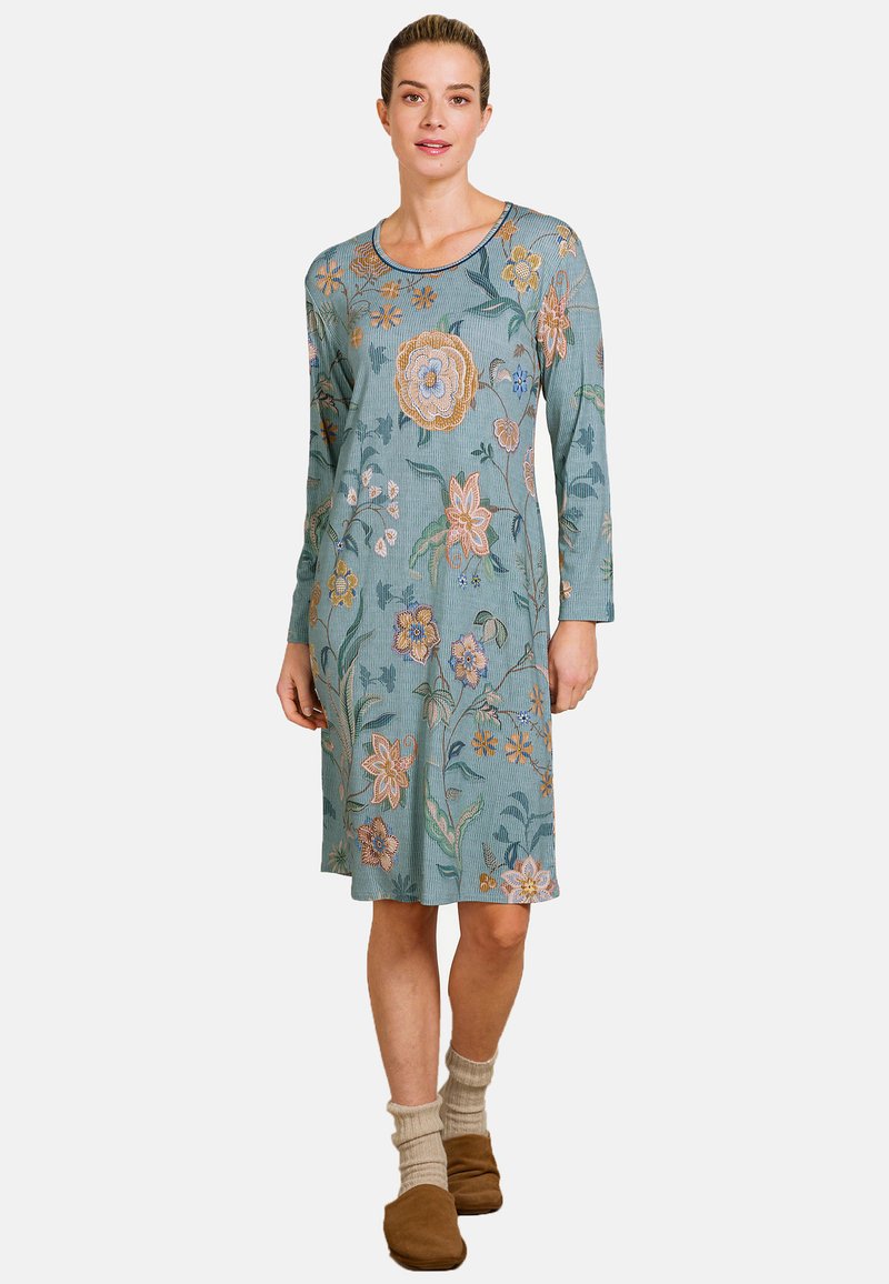 Woman wearing a knee-length, long-sleeve teal dress with multicolored floral patterns, beige socks, and brown slip-on shoes standing.