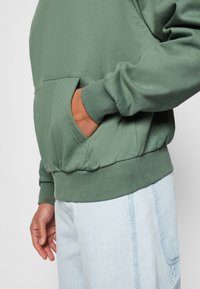 Sudadera de algodón verde con un bolsillo tipo canguro, puños y dobladillo acanalados, combinada con pantalones de color azul claro. Textura suave y corte relajado.