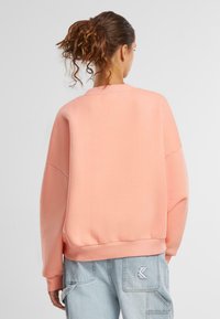 Př oversized růžová mikina s vroubkovanými manžetami a lemem. Zadní strana má hladký povrch a jednoduchý design, doplněný o světle modré džíny.
