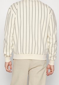 Sudadera crema con rayas verticales negras, cuello redondo, puños y dobladillo de canalé. Textura suave y ajuste holgado.