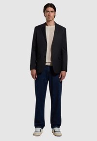 Blazer nero, maglione crema, jeans blu scuro larghi e sneakers beige. Il blazer presenta risvolti classici e due bottoni. Look casual ma curato.