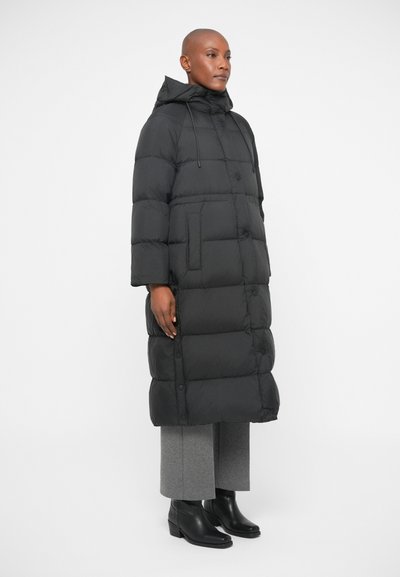 Μαύρο μπουφάν τύπου puffer με κουκούλα, με τετράγωνο σχέδιο, μεγάλες ραφές και μπροστινό κούμπωμα μ�ε κουμπιά. Φοριέται πάνω από γκρίζες παντελόνες και μαύρες μπότες.