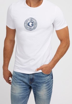 Man met een wit T-shirt met korte mouwen en een cirkelvormig blauw logo op de borst, en lichtblauwe vervaagde spijkerbroek, hand in de zak.