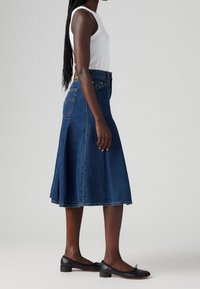 Falda en línea A de mezclilla en azul oscuro, con un bolsillo trasero y diseño plisado. Combinada con una camiseta sin mangas blanca y zapatos planos negros.