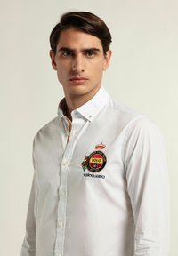 Camisa blanca de manga larga con botones, con logotipo bordado y detalle de corona. Cuenta con un cuello abotonado y sutiles acentos de color.