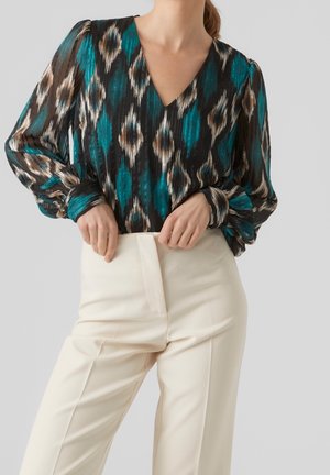 Woman wearing a long-sleeve teal, black, and beige patterned blouse with a V-neck tucked into high-waisted cream trousers.