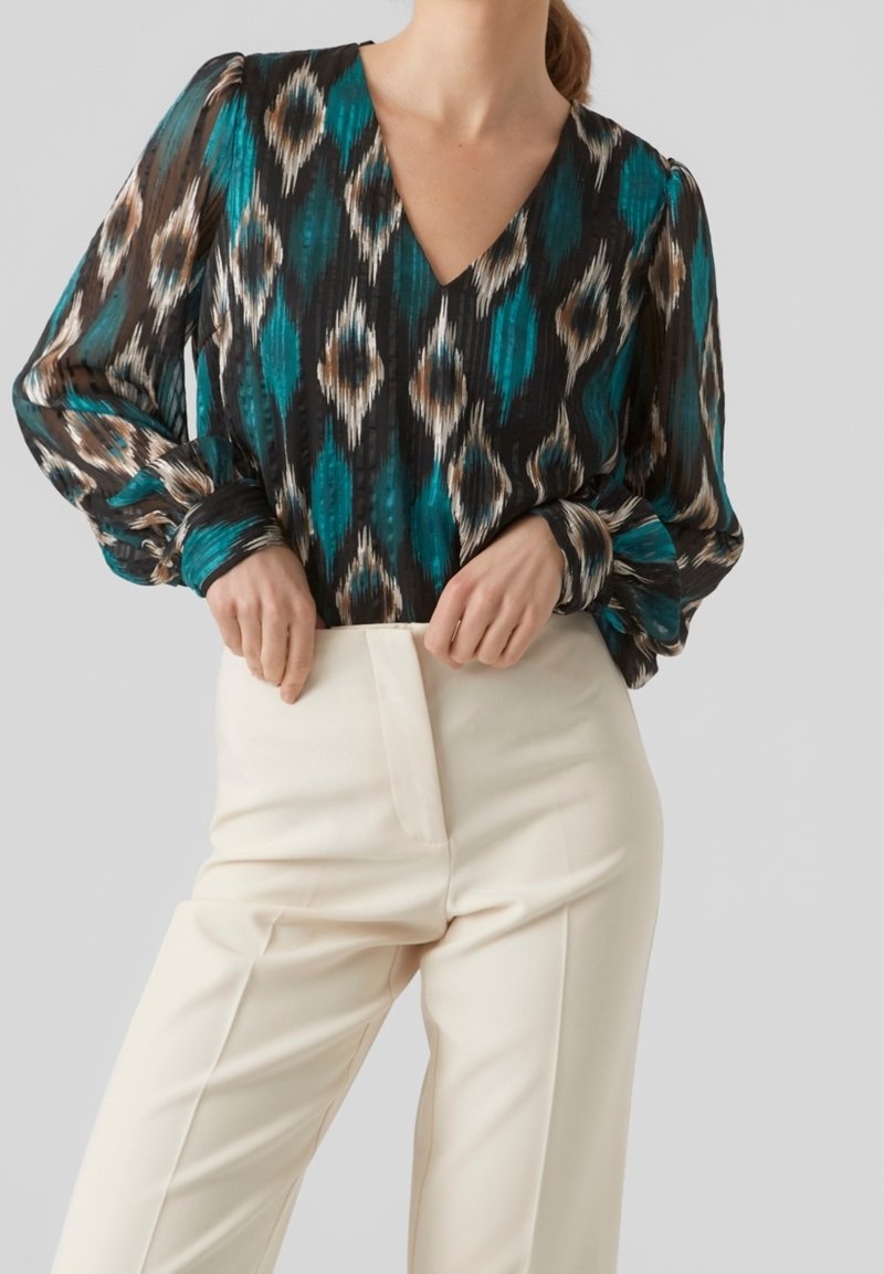 Femme portant un chemisier à manches longues à motifs bleu sarcelle, noir et beige avec un col en V, rentré dans un pantalon crème taille haute.