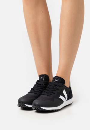 Jambes portant des baskets noires et blanches à lacets avec un logo V blanc sur les côtés, sur un fond uni.