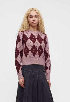 Jeune femme aux longs cheveux blonds bouclés portant un cardigan à losanges rose et bordeaux et une jupe plissée foncée.