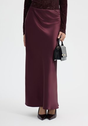 Donna che indossa una gonna lunga e liscia bordeaux, scarpe appuntite bordeaux e tiene una piccola borsa nera al suo fianco.