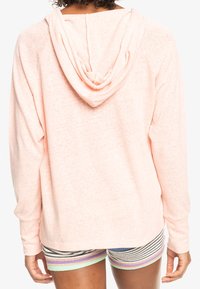 Persona che indossa un felpa con cappuccio rosa chiaro e pantaloni corti strizzati multicolori, mostrata di spalle su uno sfondo bianco.