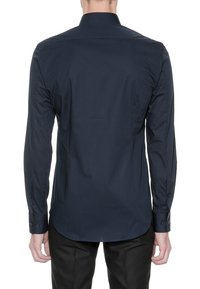 Homme portant une chemise habillée à manches longues bleu marine foncé et un pantalon noir, vu de dos sur fond blanc.