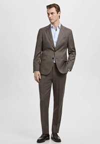 Rjav krojen kostim z enojno gumbo jopiča, dvema sprednjima žepoma in ujemajočimi se hlačami. Svetlo modra srajca in črni čevlji.