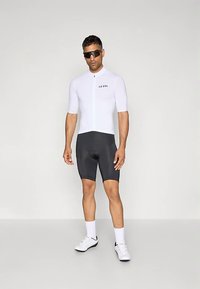 Hvit sykkeltrøye med korte ermer og glidelås foran, med svart logo, kombinert med svarte sykkelshorts. Modellen har på seg solbriller og hvite sykkelsko.