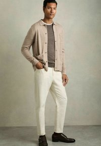 Cárdigan beige con botones sobre un suéter marrón, acompañado de pantalones blancos y zapatos marrones tipo mocasín. Texturas suaves destacadas y diseño ajustado.
