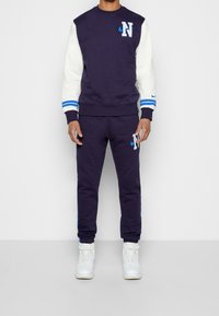 Mörkblå sweatshirt med vita ärmar, med en "N"-logotyp och blåa detaljer. Matchad med mörkblå sweatpants, båda i mjukt material, och vita sneakers.