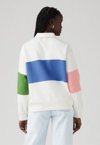 Balta kolārpārcēnā ar horizontālām svītrām: zaļa, zila un rozā. Ar garām piedurknēm un ribotām aprocēm un apakšmalu.