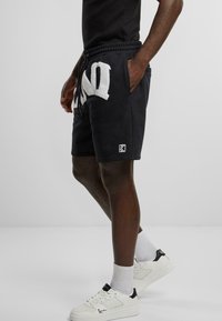 Black shorts with white plush lettering "NO" on the front, elastic waistband with drawstring, and a small logo patch at the hem.