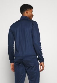 Sötétkék sportdzseki magas gallérral, hosszú ujjaival és fehér logó részletekkel az ujjak mentén. Simább, lélegző anyagból készült.