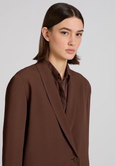 Frau mit glattem, schulterlangem braunem Haar, die ein dunkelbraunes Sakko und ein dazu passendes Hemd mit Knöpfen vor einem einfarbigen Hintergrund trägt.