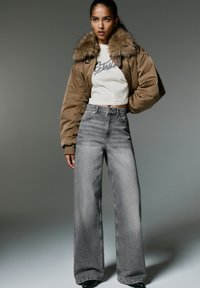 Jachetă bomber maro cu guler din blană, asociată cu blugi gri din denim cu talie înaltă și picioare largi, și un tricou scurt alb cu imprimeu grafic.