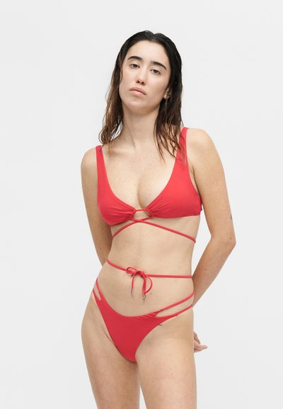 Γυναίκα με βρεγμένα μαλλιά που φοράει κόκκινο μπικίνι με λουριά που τυλίγονται γύρω, στέκεται μπροστά από απλό λευκό φόντο και κοιτάζει την κάμερα.